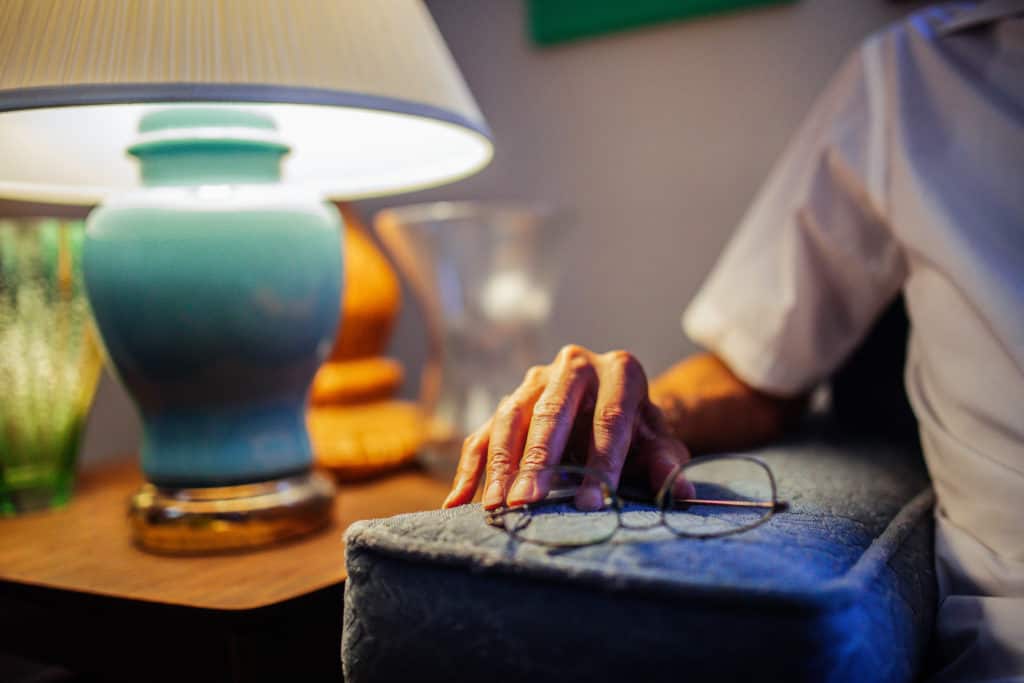 Elderly man's hand rests on the arm of his couch, next to glasses