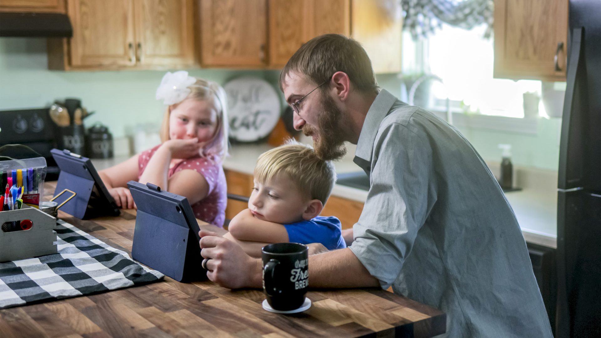 Father helping two children with tablets at kitchen island